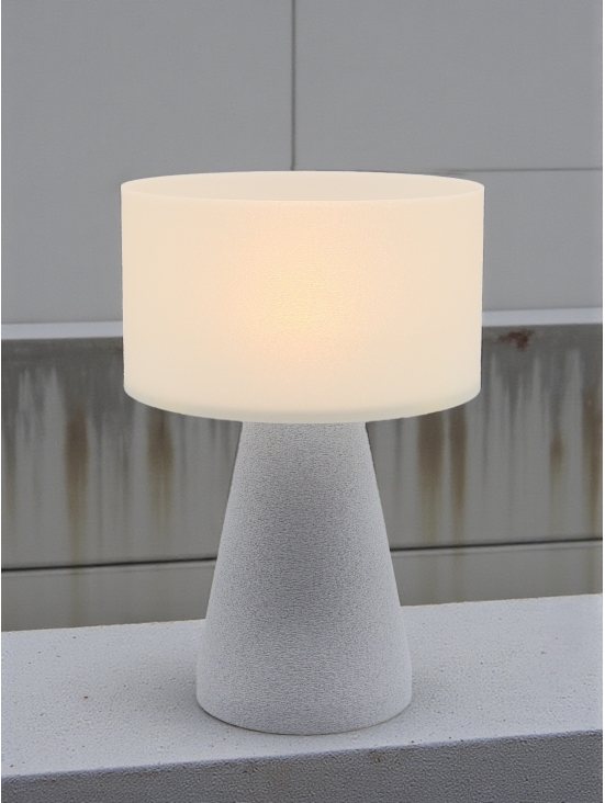 Lampe à poser design Meredith granit pierre allumée, lumière douce, ambiance industrielle sur béton et mur gris.